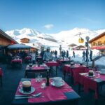 A panoramic, photorealistic winter scene of a bustling ski resort in the German Alps, shot in 16:9 format. In the foreground, a neatly set outdoor dining terrace of a mountain restaurant features empty tables with red-checkered cloths, steaming coffee cups, and lit candles, suggesting a peak season atmosphere. The background shows snow-covered slopes, a ski lift, and guests in winter gear arriving and departing. The lighting is soft natural daylight with clear blue sky, emphasizing a dynamic yet inviting hospitality environment. No text, logos, or visible faces are present. The image conveys the high demand for service staff in seasonal tourism.
